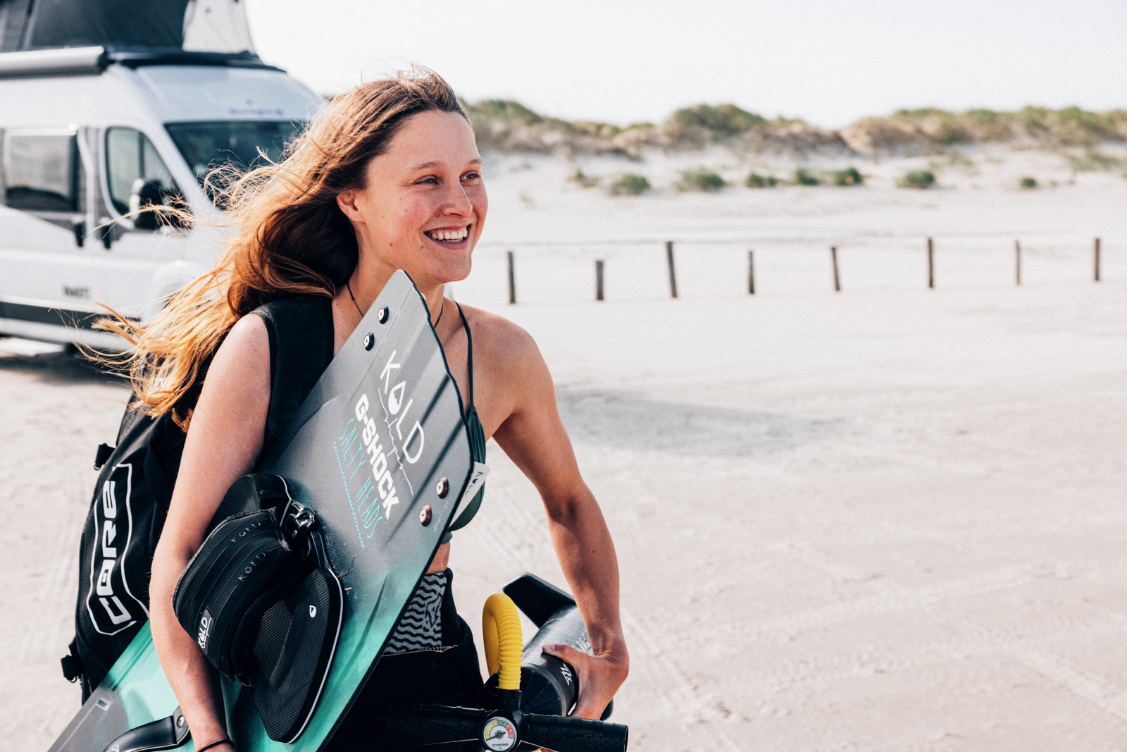A traveler carrying watersports gear by the beach with a campervan behind her