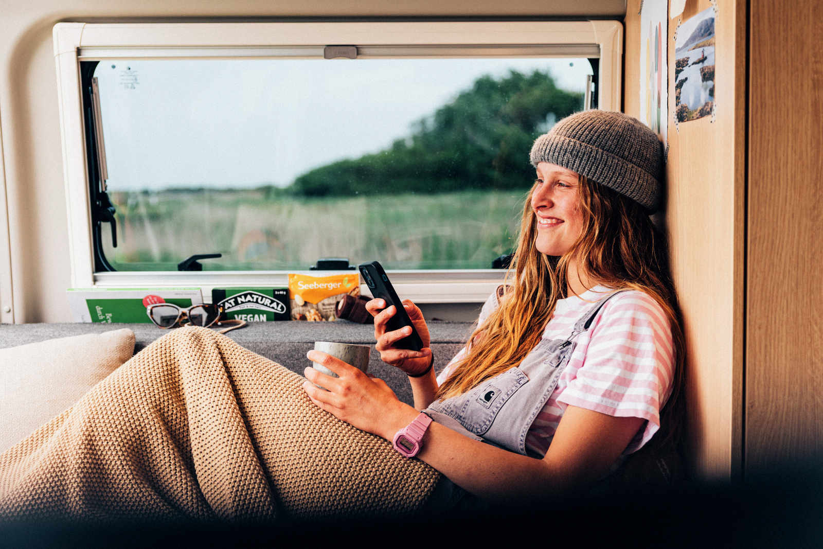 A traveler relaxing under a blanket in a campervan with a phone and a cup
